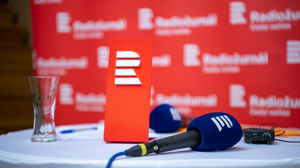 Detailní záběr na mikrofony, sklenici a podium s logem RenéNEKUDA.cz na pozadí s nápisem Radiózní České mojí.