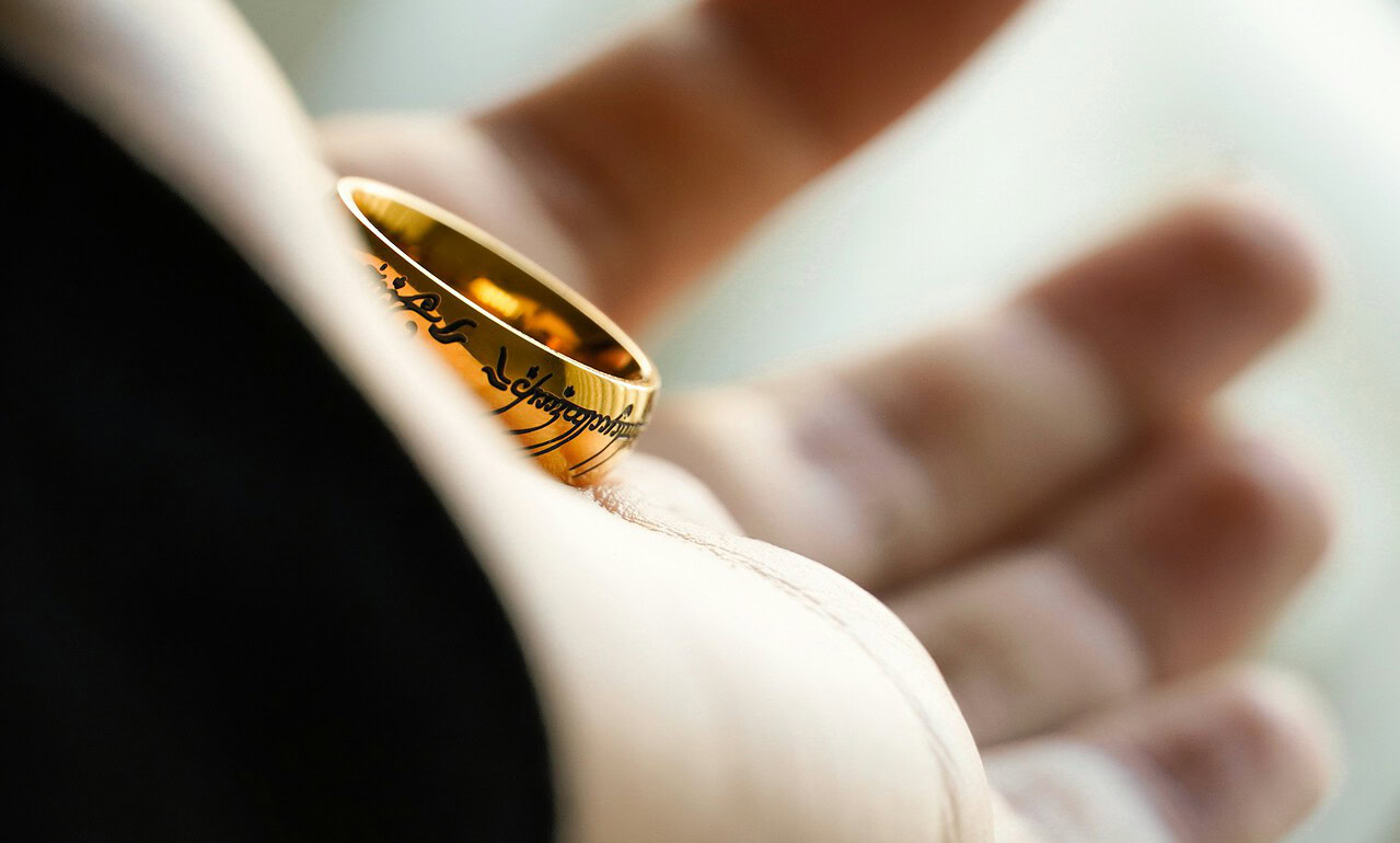 Záběr na zlatý prsten s nápisem, který je umístěn na prstě osoby, symbolizující spojení a závazek.
